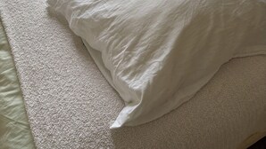 Ein ordentlich bezogenes Bett mit weißer Decke und Kissen, ein Nachttisch und ein Fenster mit Vorhängen.