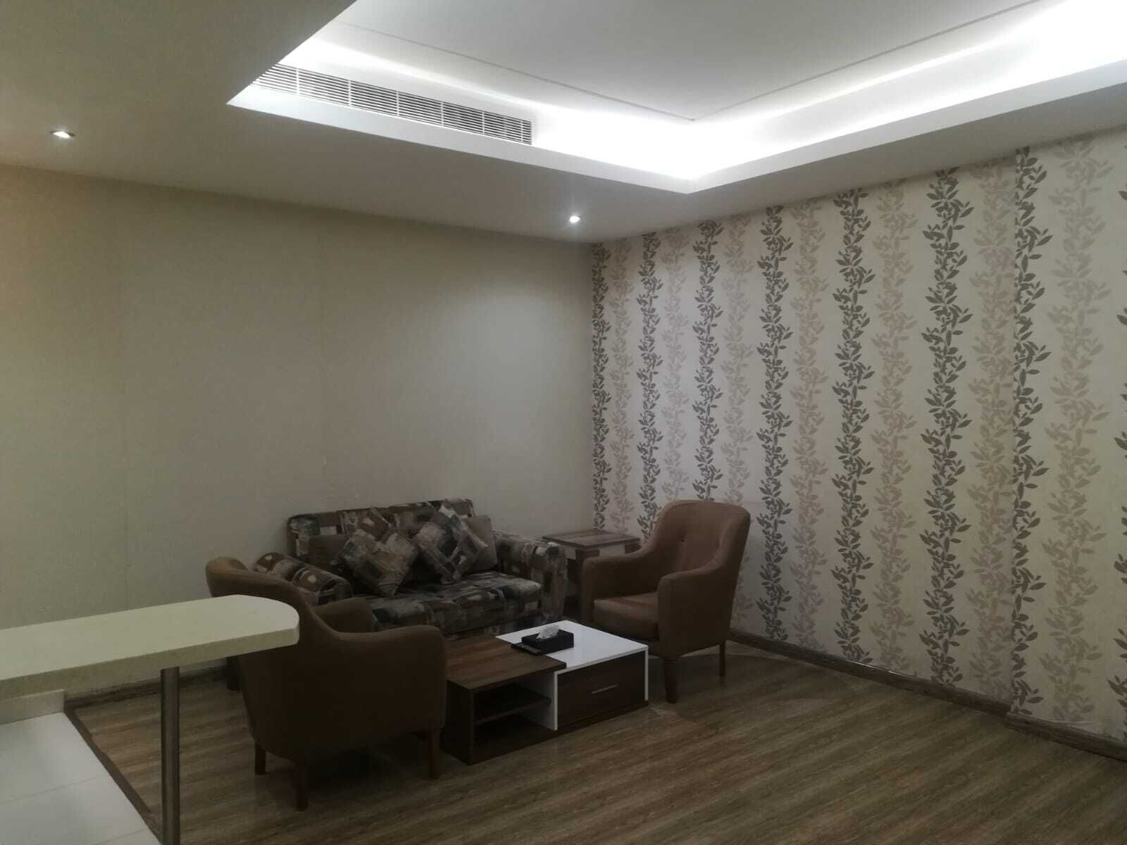 Ein Wohnzimmer mit einer Couch, zwei Sesseln, einem Couchtisch und einem kleinen Tisch. Die Wände sind mit ornamentalem Papier tapeziert und der Boden ist aus Holz.