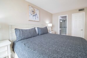 A bedroom with a large bed, a nightstand, a lamp, a bathroom door, and a framed picture on the wall. - Bubbles Up ~ Beachfront & Top Corner Balcony (Folly Beach)