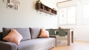 A modern living room with a grey sofa, two cushions, a wooden shelf with plants, and a dining area with chairs and a table. - Newly renovated apartment, located in the lively Bicocca area, right in front of the Arcimboldi Theatre, which offers shows and concerts throughout the year.The location is ideal: a stone's throw from the Greco train stop, from which you can reach the  (Milano)