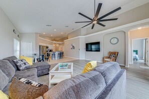 Ein geräumiges Wohnzimmer mit einer grauen Sofagarnitur, einem Couchtisch und einem Deckenventilator. Im Hintergrund sind eine Küche und ein Essbereich zu sehen.