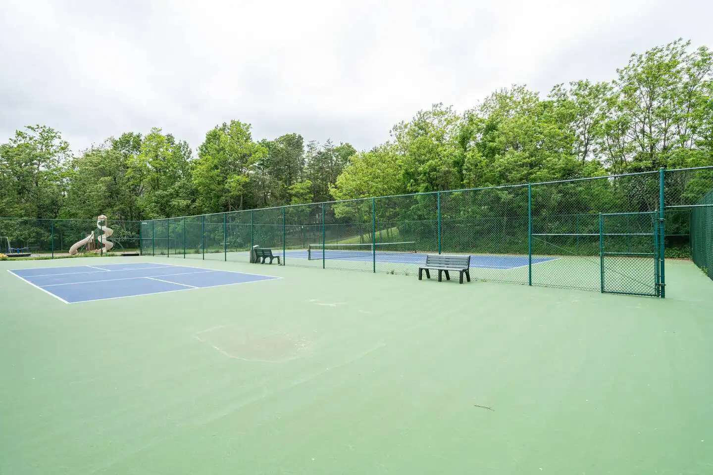 En udendørs tennisbane med grøn overflade, omgivet af et grønt hegn og træer.