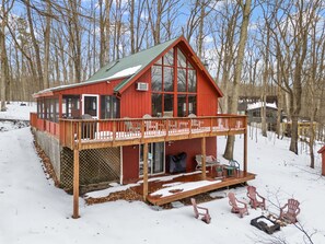 Eine rote Holzhütte mit einer Holzterrasse und einem großen Fenster, umgeben von Bäumen und Schnee.