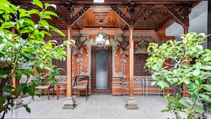 Una estructura de madera ornamentada con una puerta central, flanqueada por dos sillas y una mesa, rodeada de follaje.