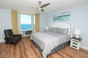 Ein Schlafzimmer mit Bett, Sessel, Nachttisch, Lampe und Meerblick.