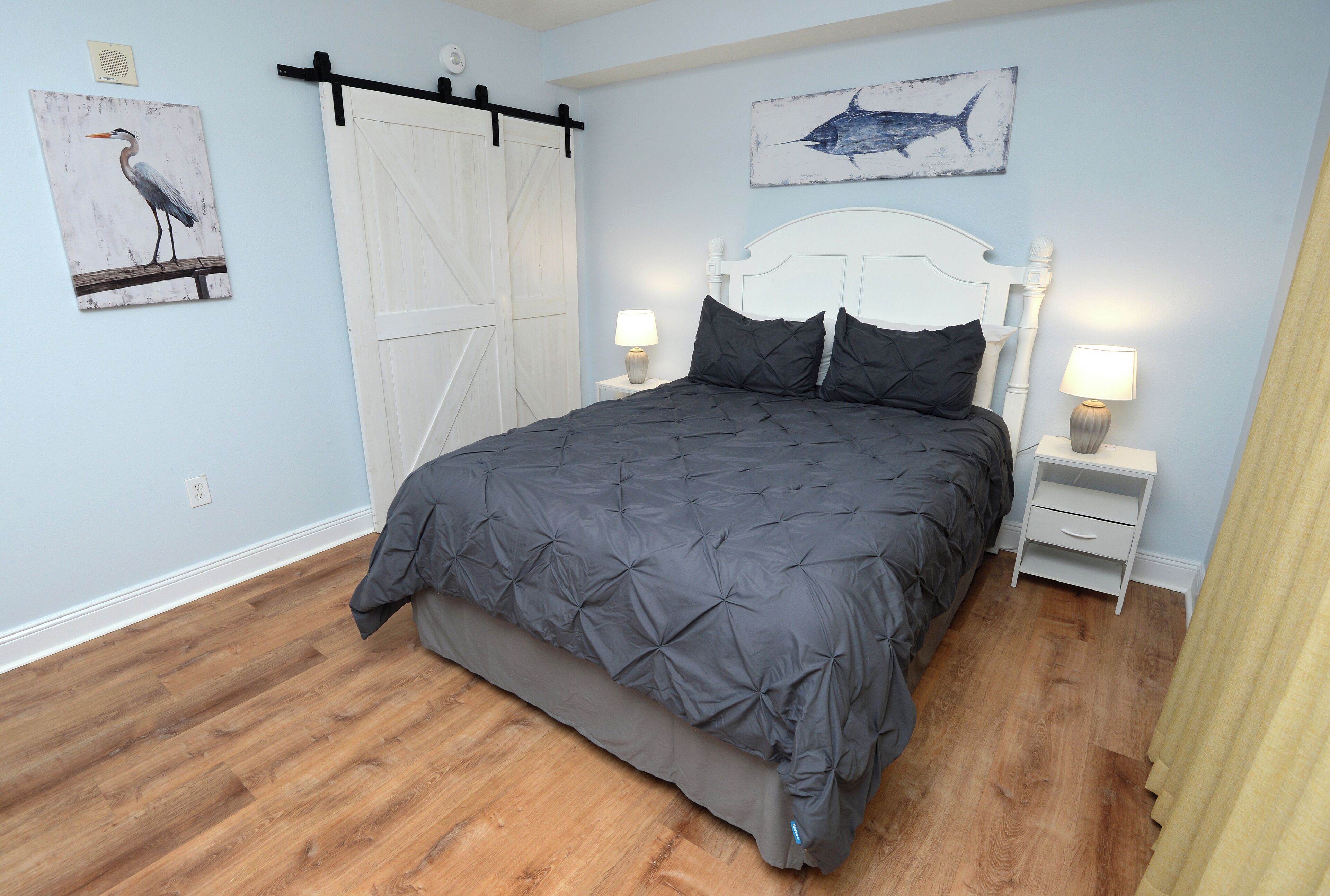 A bedroom with a bed, two bedside tables, a nightstand, a white headboard, a wooden floor, a wall-mounted artwork of a bird, and a wall-mounted artwork of a shark.