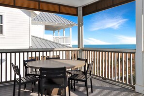 A covered outdoor area with a round table and chairs, a white building with a balcony, and a view of the ocean. - Southern Charm (Port St. Joe)