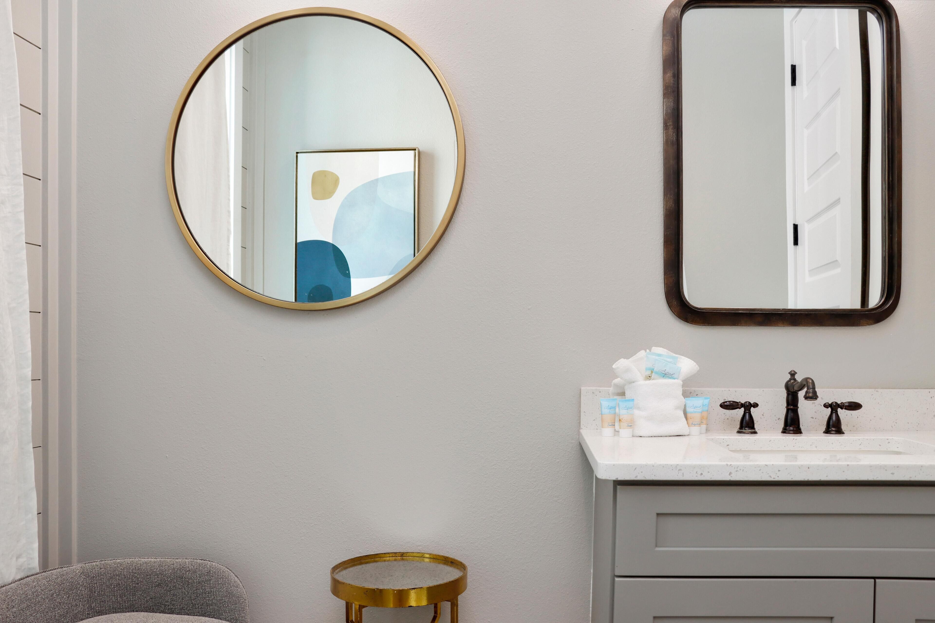 Een badkamer met een ronde spiegel, een rechthoekige spiegel, een wastafel met twee kraan en een klein rond krukje.