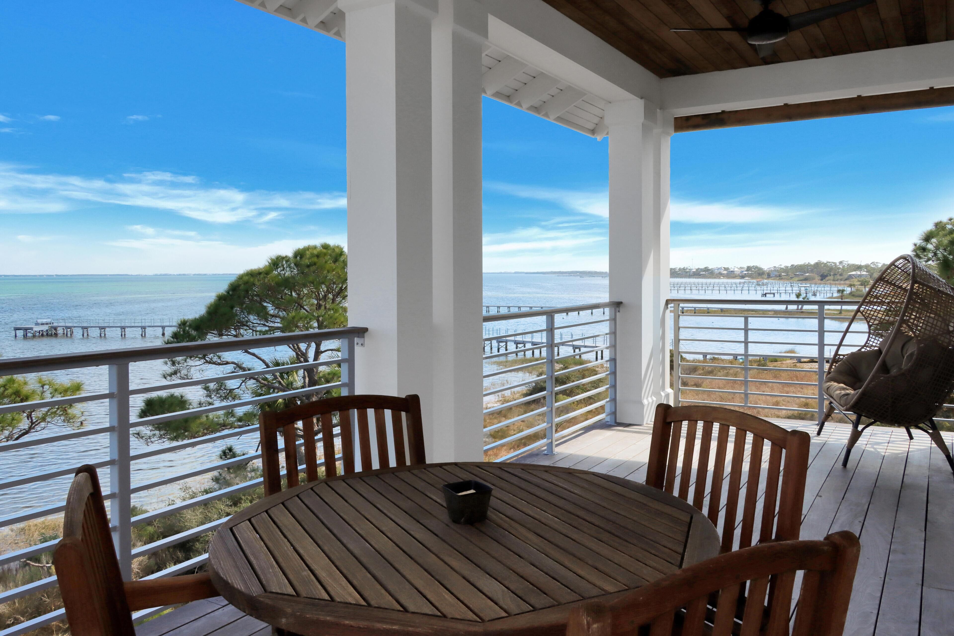 A wooden deck with a table and chairs, overlooking a body of water with a pier and trees.