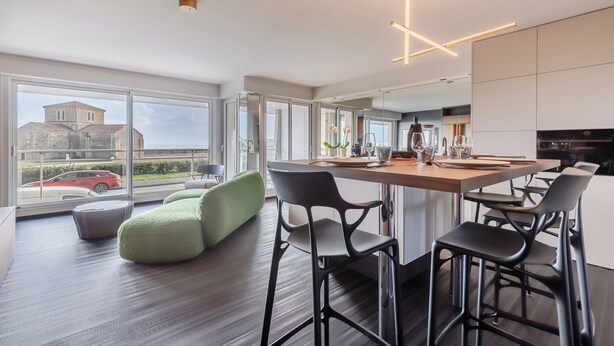 Cocina moderna con una isla central, taburetes de bar y ventanas amplias que ofrecen vista a un edificio y un coche rojo afuera.