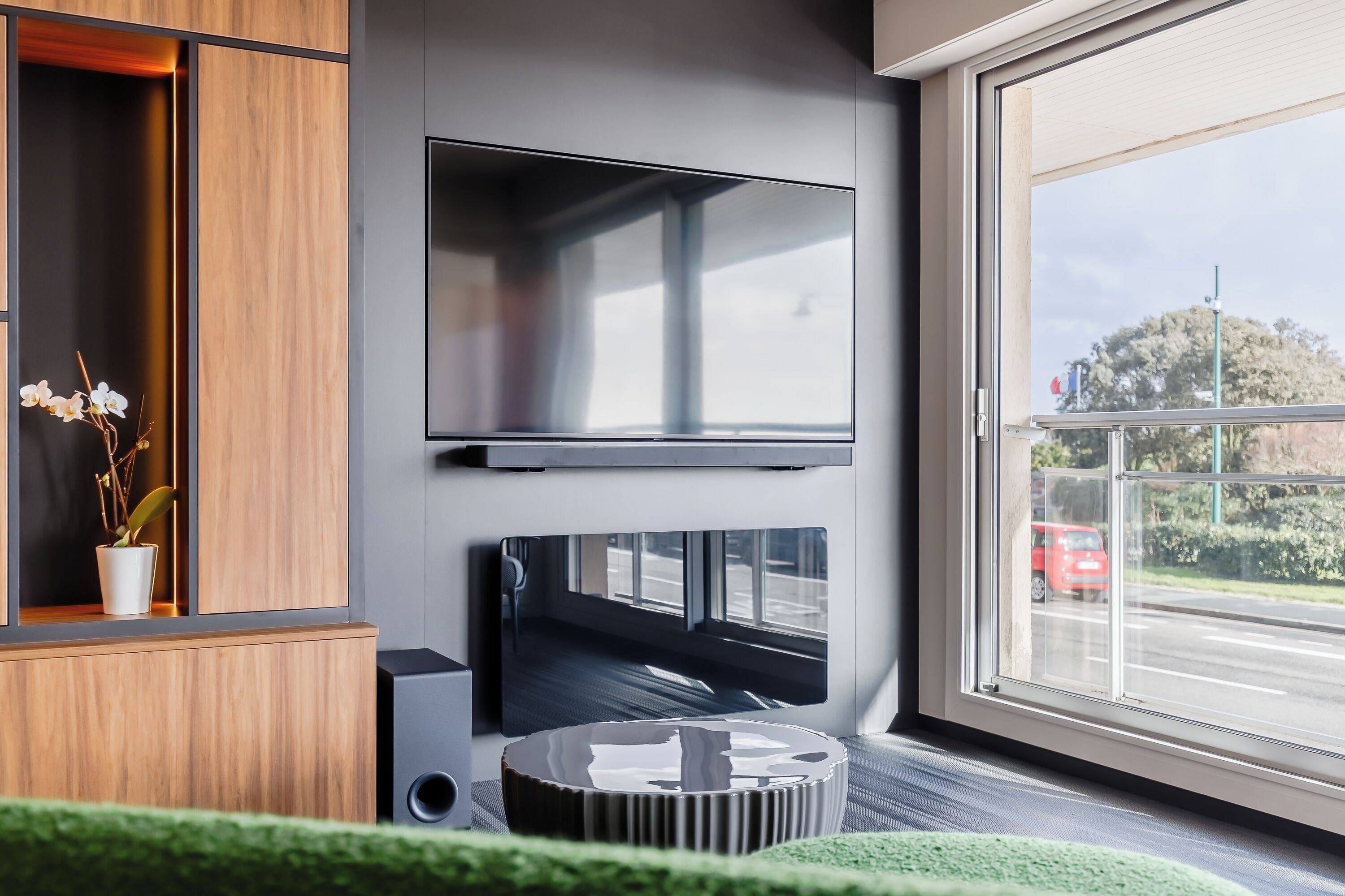 A modern living room with a flat-screen TV mounted on the wall, a wooden cabinet with a plant, and a view of the street through a large window.