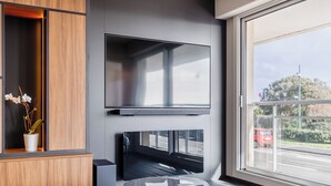 A modern living room with a flat-screen TV mounted on the wall, a wooden cabinet with a plant, and a view of the street through a large window. - L'Écrin des Sables 4*, an exceptional 70 m2 seafront location (Les Sables-d'Olonne)