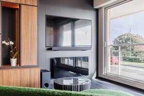 A modern living room with a flat-screen TV mounted on the wall, a wooden cabinet with a plant, and a view of the street through a large window. - L'Écrin des Sables 4*, an exceptional 70 m2 seafront location (Les Sables-d'Olonne)