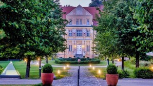 Um edifício grande e imponente, com fachada simétrica, entrada central e um jardim bem cuidado com um caminho pavimentado que leva à entrada.