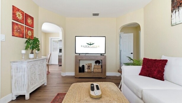 Een woonkamer met een open haard, een televisie erboven en een witte bank met een rode sierkussen. Links staat een witte kast met decoratieve objecten en rechts een plant.
