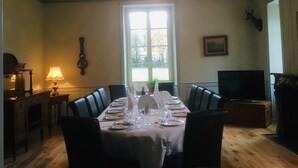 Une salle à manger avec une longue table dressée pour six personnes, un lustre, un téléviseur et une tête de cerf décorative accrochée au mur.