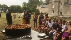 Un groupe de personnes s’est rassemblé autour d’un grand barbecue installé à l’extérieur, près de nourriture, dans un espace verdoyant avec un bâtiment en pierre en arrière-plan.