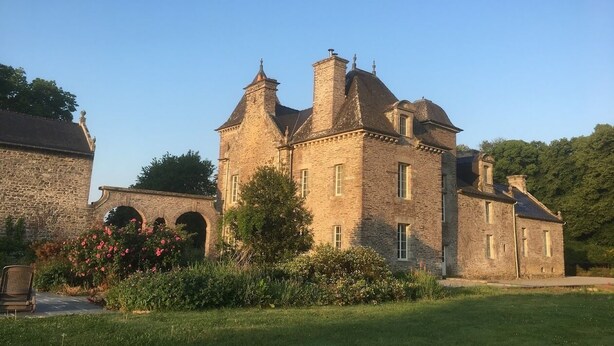 Un grand bâtiment en pierre, doté de nombreuses cheminées et de fenêtres en arc de cercle, entouré d’un jardin bien entretenu où poussent des plantes à fleurs.
