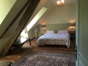 Un dormitorio con techo inclinado, una cama con ropa de cama floral, un banco de madera, un ventilador de pie y una alfombra estampada.