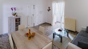 Ett modernt vardagsrum med ett matbord i trä, ett soffbord i glas och en grå soffa. Det finns en vit dörr och ett fönster med tunna, genomskinliga gardiner.