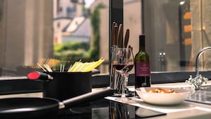 Un comptoir de cuisine avec une poêle, un verre de vin rouge, une bouteille de vin, un bol de tomates cerises et une fenêtre donnant sur des immeubles.