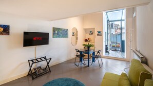Un salon moderne doté d’un téléviseur à écran plat, d’un coin repas avec une table et des chaises, d’un canapé vert et d’une vue sur un balcon extérieur, accessible par une porte vitrée.