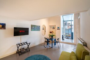Ein modernes Wohnzimmer mit Flachbildfernseher, Essbereich mit Tisch und Stühlen, einem grünen Sofa und Blick durch eine Glastür auf einen Balkon.