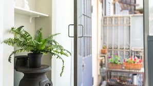 Une fougère en pot posée sur un piédestal, une étagère blanche ornée d’une petite statue et, au fond, une grille en métal agrémentée d’une clôture décorative.
