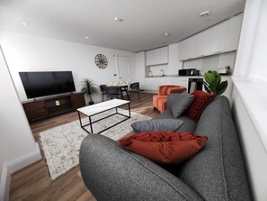 Un salon moderne avec un canapé, une table basse et un téléviseur. On y trouve également un coin repas avec des chaises et une table. La cuisine est visible, avec ses placards et ses appareils électroménagers.