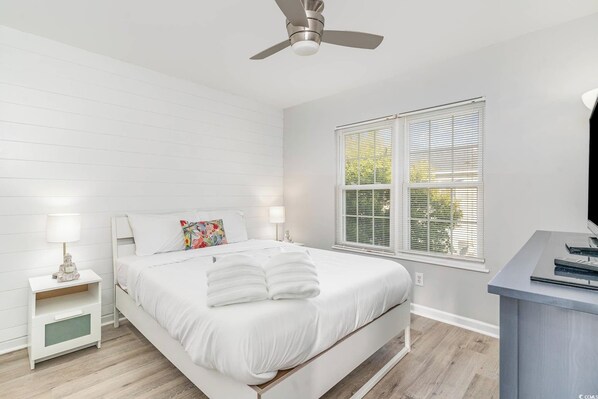 A bedroom with a bed, nightstands, a ceiling fan, a window with blinds, and a desk with a chair. - Walk to Beach, Pool, King Suite, Sleeps 8 (North Myrtle Beach)