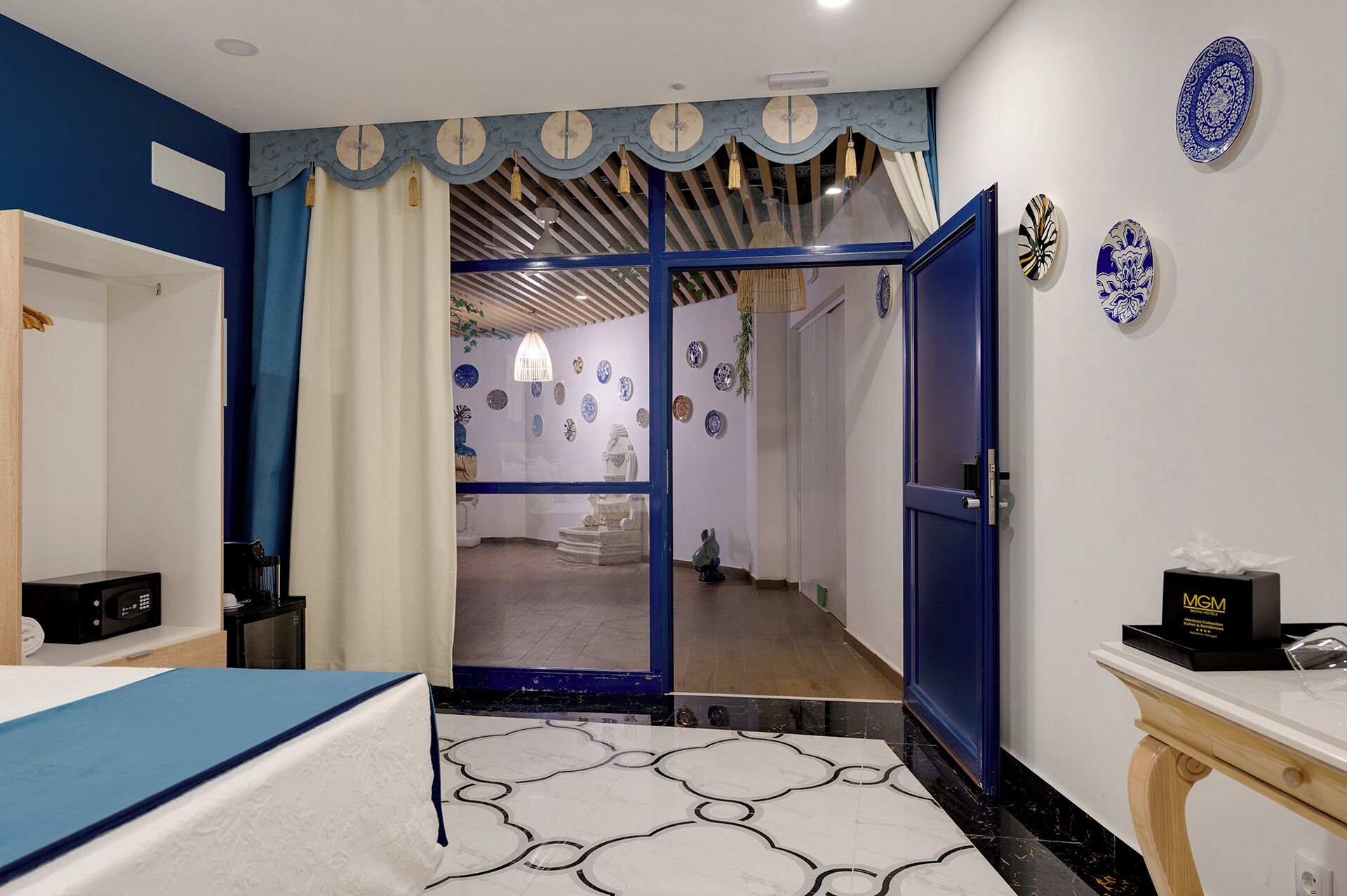 A hotel room with a bed, a desk, a blue door, and a hallway leading to a bathroom.