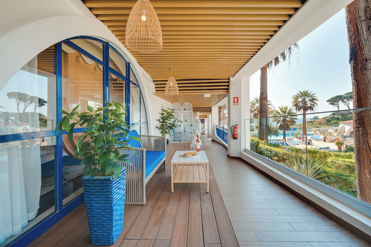 A corridor with a wooden ceiling, a seating area with a table, and a view of a pool and palm trees.