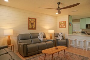 A living room with a sofa, two armchairs, a coffee table, and a ceiling fan. There are two lamps on side tables, a rug on the floor, and a kitchen area in the background. - Sea-turtle (St. Augustine)
