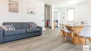 Una sala de estar moderna con un sofá gris, una mesa de comedor de madera con sillas y una cocina al fondo.