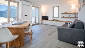 Un moderno salón con un sofá gris, una mesa de comedor de madera con sillas, un televisor y un balcón con vistas a la montaña.