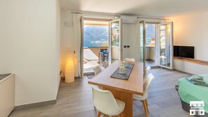 Una sala de estar moderna con una mesa de comedor de madera, sillas blancas, un televisor y un balcón con vistas a la montaña.