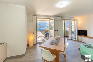 Ein modernes Wohnzimmer mit Esstisch aus Holz, weißen Stühlen, einem Fernseher und einem Balkon mit Blick auf die Berge.