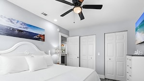 Une chambre à coucher avec un lit, un ventilateur de plafond, un tableau représentant une plage et une armoire.