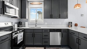 Une cuisine moderne avec des meubles de couleur sombre, des appareils en acier inoxydable, une fenêtre avec des stores et des luminaires suspendus.