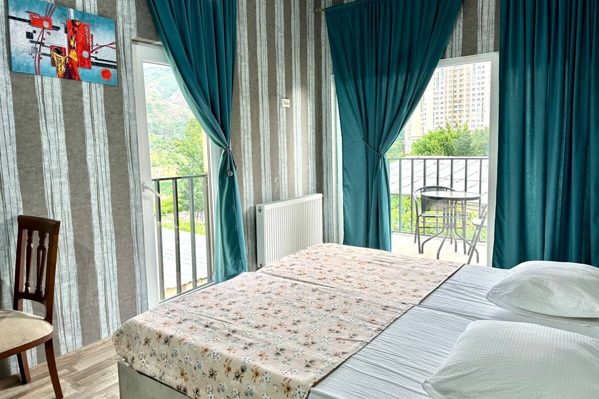 A bedroom with a bed, a chair, a view of the outdoors, and a painting on the wall.