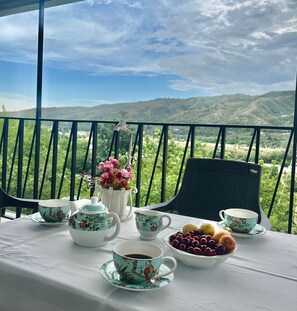 A table set with tea cups, a teapot, a vase with flowers, and a bowl of fruit on a balcony with a mountain view. - Holiday Garden Tbilisi (Tbilisi)