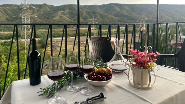 A table set with wine glasses, a bottle, a bowl of fruit, and a vase of flowers on a balcony with a mountain view. - Holiday Garden Tbilisi (Tbilisi)