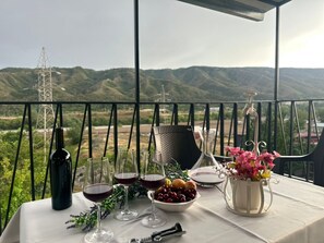 Une table dressée avec des verres à vin, une bouteille, un saladier de fruits et un vase de fleurs, le tout sur un balcon offrant une vue sur la montagne.