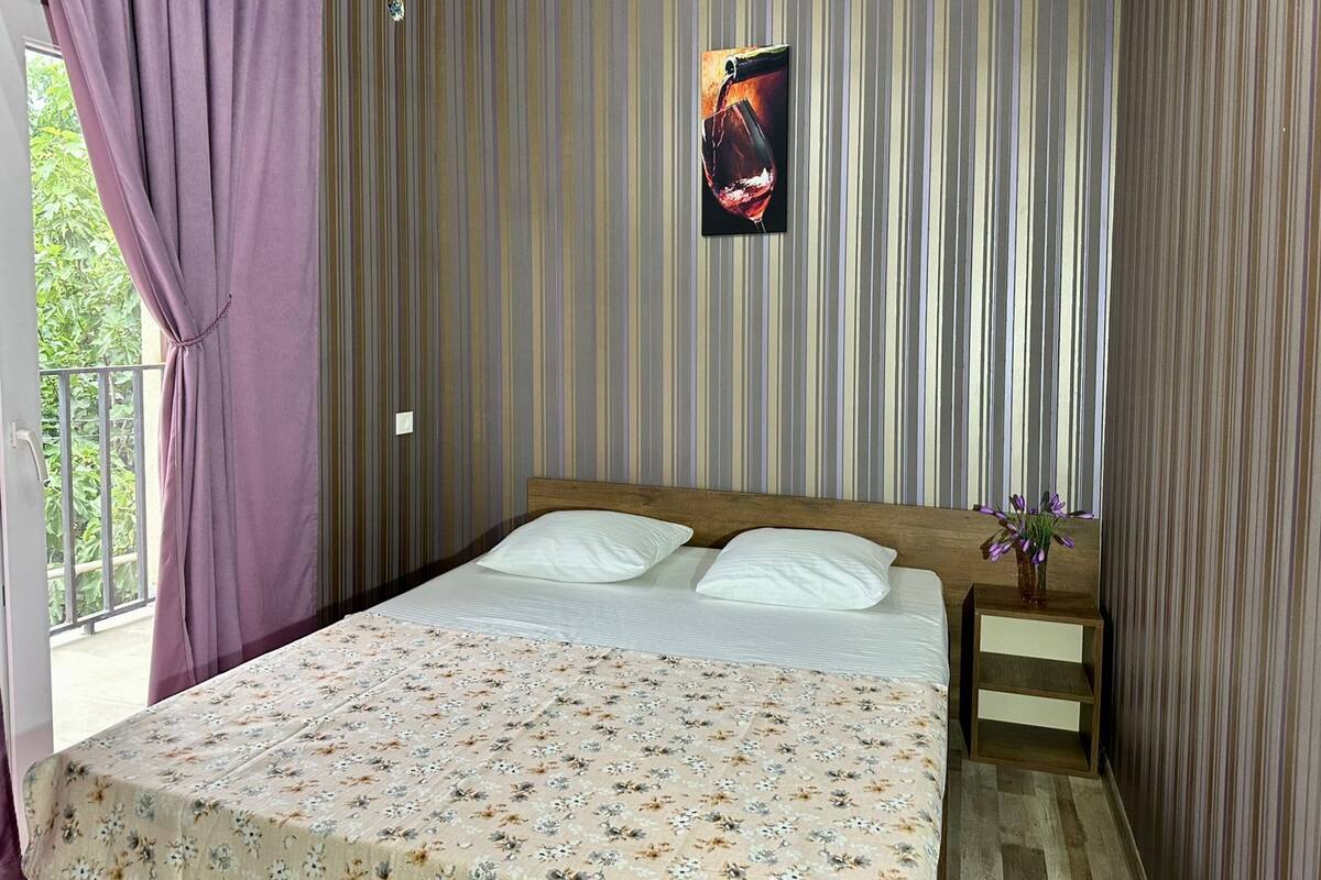 A bedroom with a bed, a nightstand, a wall-mounted picture, and a window with curtains.