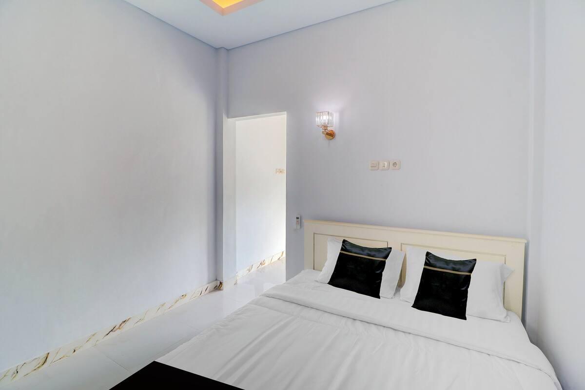 A neatly made bed with white bedding and two black pillows, a bedside lamp, and a door leading to another room.