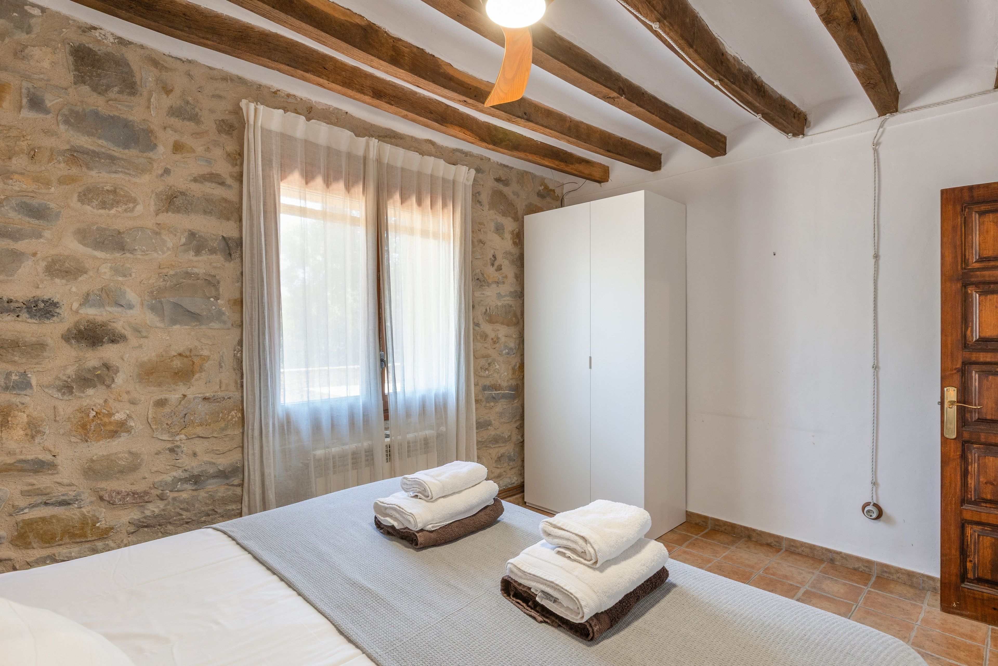 A bedroom with stone walls, a wooden ceiling, a bed with a white mattress and grey blanket, two folded towels, a white wardrobe, and a wooden door.