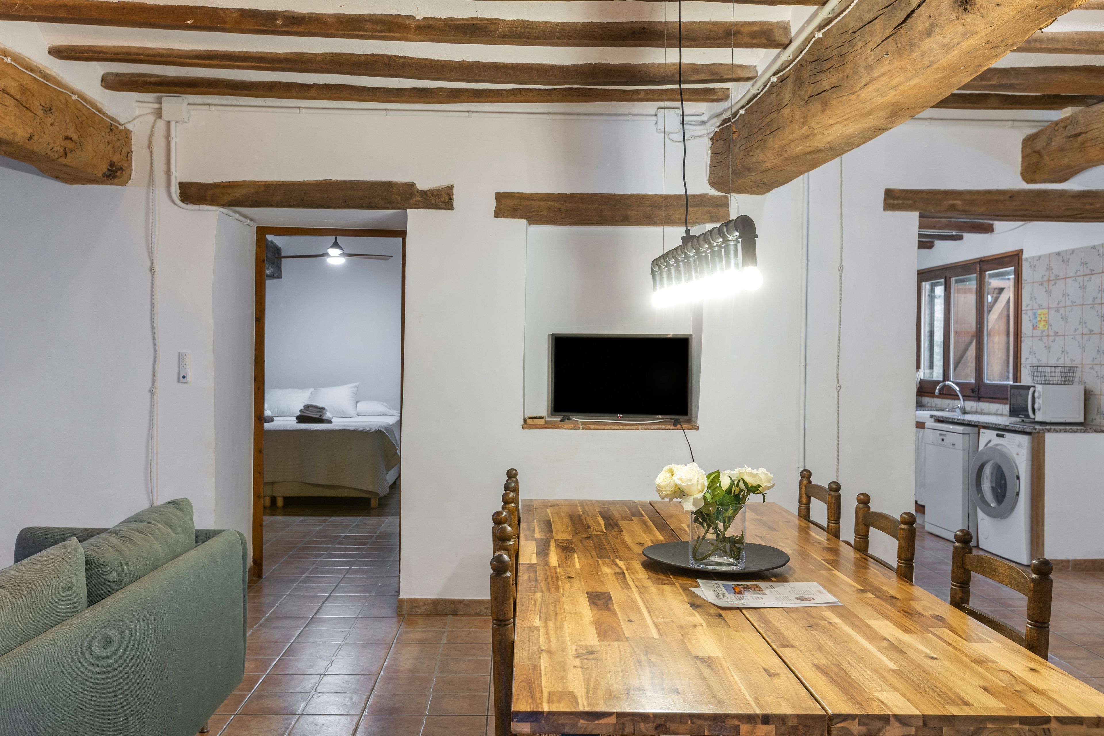 Una habitación con una mesa de comedor de madera, sillas, un televisor montado en la pared y una lavadora en la esquina.