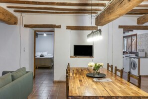 Ein Zimmer mit Esstisch aus Holz, Stühlen, einem an der Wand befestigten Fernseher und einer Waschmaschine in der Ecke.