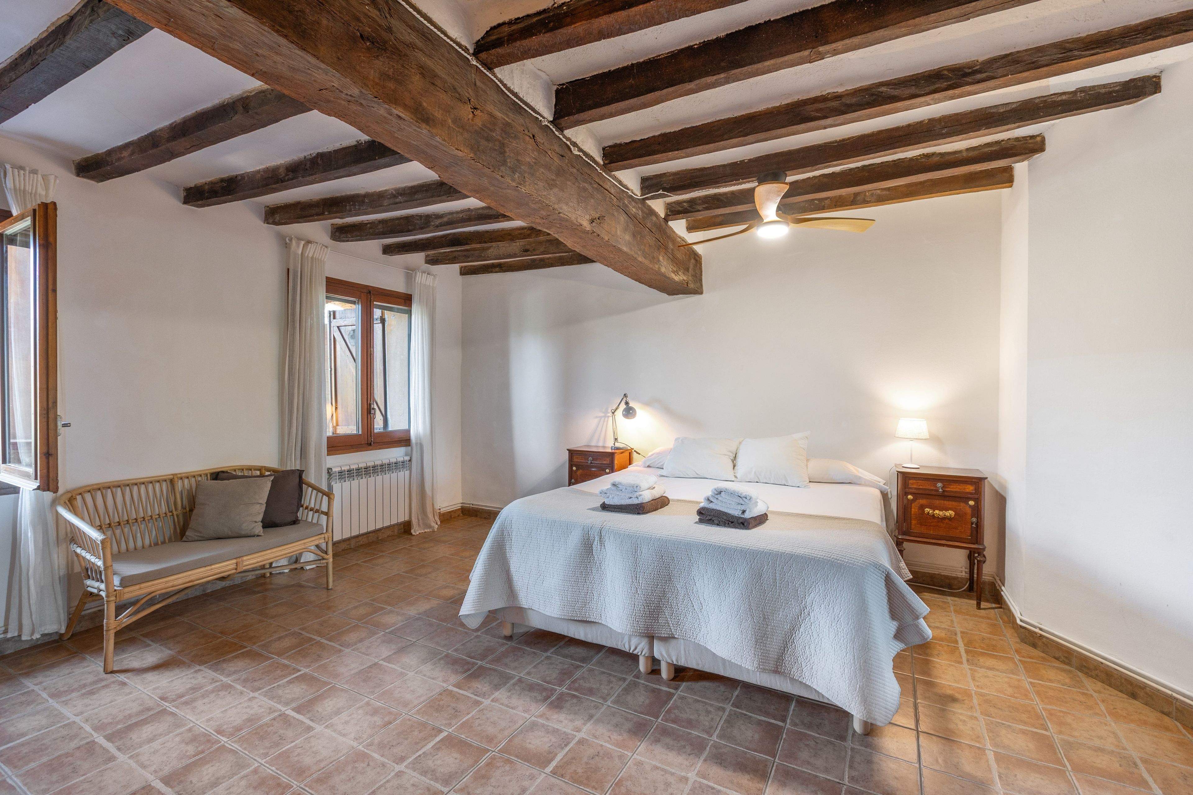 Ein Schlafzimmer mit Bett, Nachttischen, einem Fenster mit Vorhängen, einer Holzbalkendecke und einem Fliesenboden.
