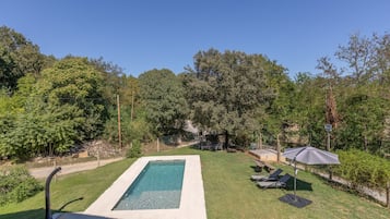 Una piscina rectangular con solárium empedrado, rodeada de césped y árboles.